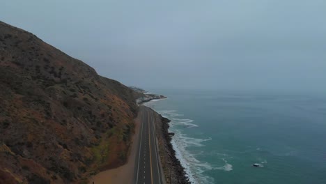 slow pull back establishing drone shot as car pulls out onto pacific highway one, near malibu on a grey and foggy morning