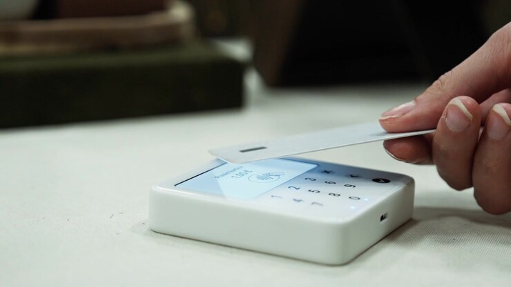 A hand holds a payment card over a small wireless reader on a table. The screen processes the contactless transaction and displays a confirmation.