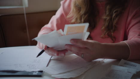 woman in pink sweater sifts through receipts at kitchen table in daytime, marking down totals and organizing finances