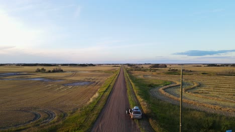 Backwards-flying-drone-revealing-silver-minivan-parked-along-a-long-and-endless-dirt-road-in-flagstaff-county,-Alberta