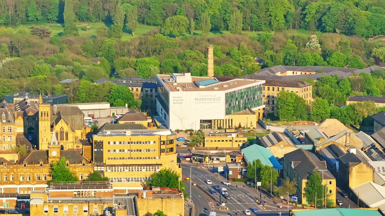 Drone shot reveals University of Huddersfield campus featuring the modern Oastler Building, historic brick mills, surrounding streets, and lush tree areas in the background, aerial view, drone shot