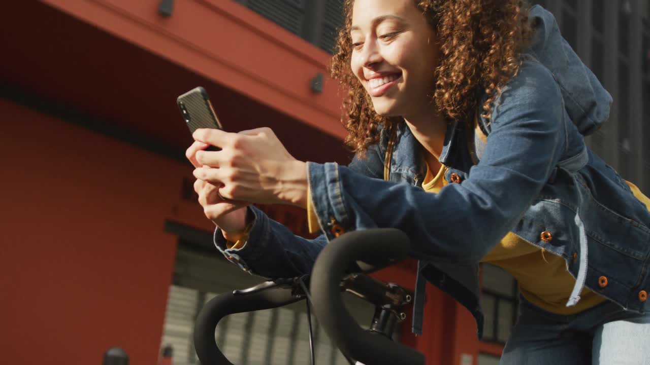 Happy biracial woman in city, sitting on bike using smartphone