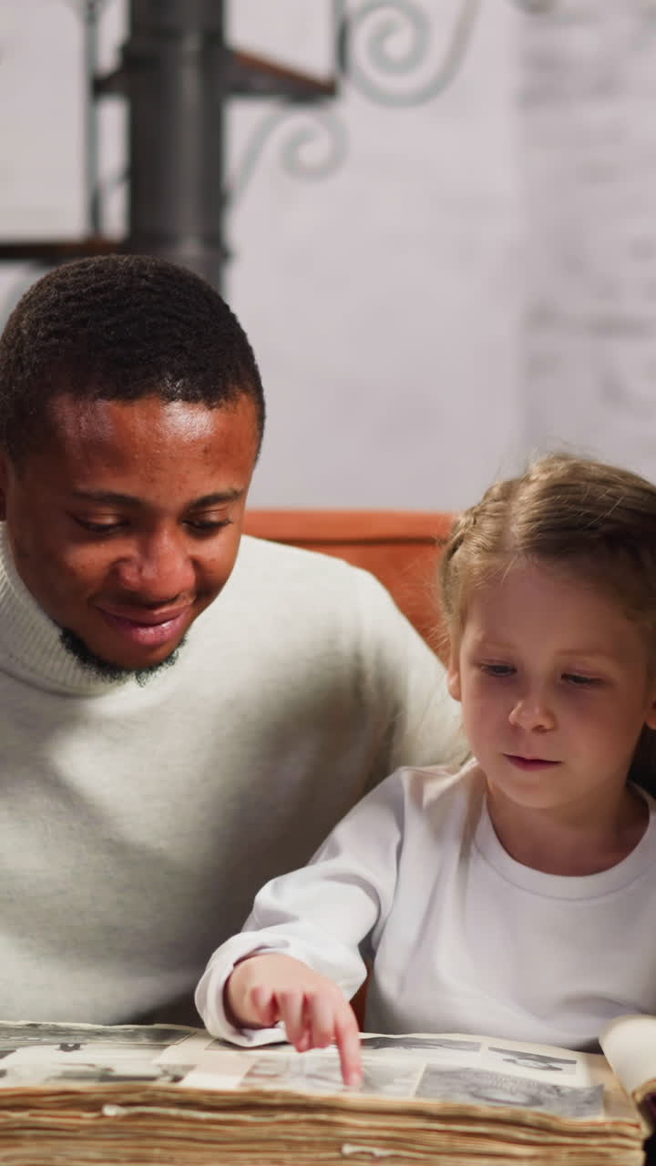 Preschooler girl steps by fingers on page of vintage family photo album page sitting between African-American father and white mother at home slow motion