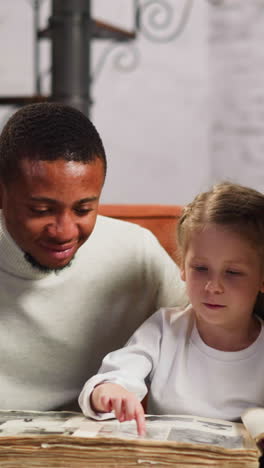 preschooler girl steps by fingers on page of vintage family photo album page sitting between african-american father and white mother at home slow motion