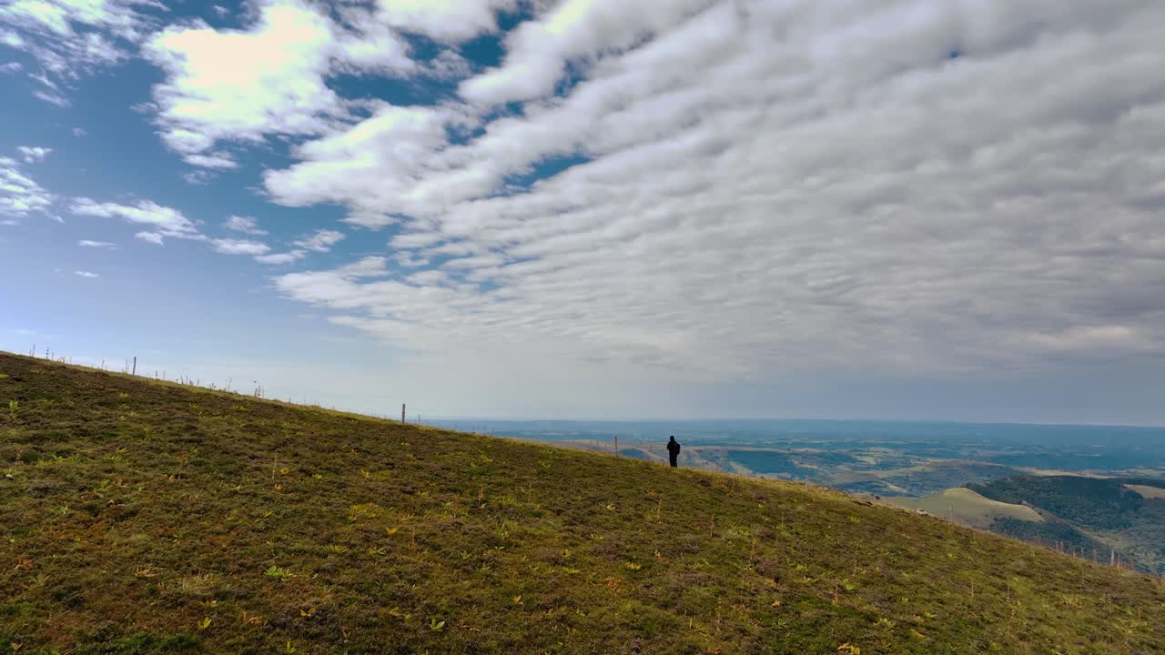 Perspectiva aérea de dron mostrando a un fotógrafo solitario de pie en una amplia ladera cubierta de hierba. Vasto paisaje bajo un cielo nuboso texturizado