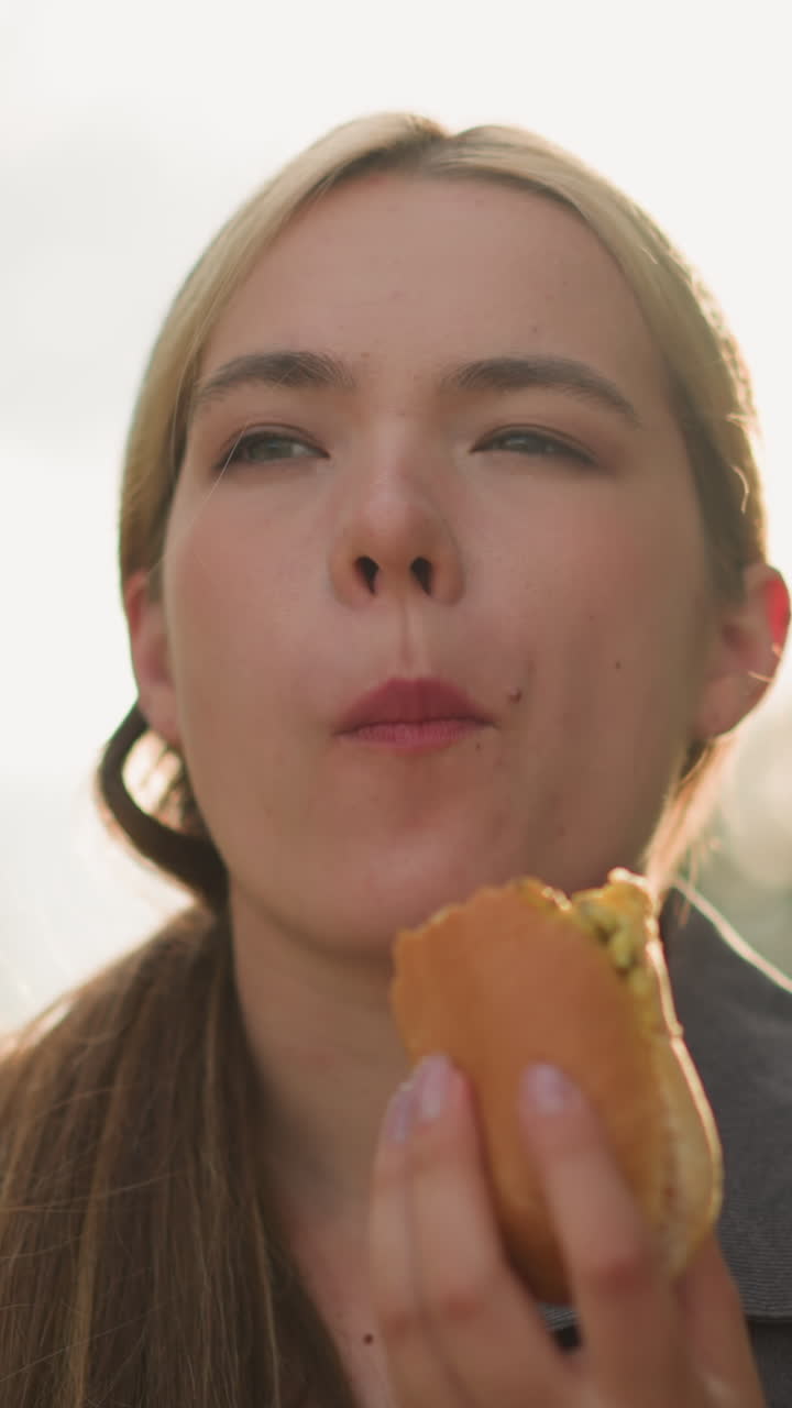 dama con camiseta gris disfruta de comer hamburguesa con una expresión reflexiva en un entorno al aire libre iluminado por el sol, luz suave iluminando su cara, el fondo está borroso con los cálidos rayos del sol