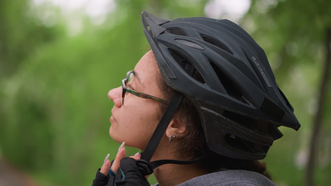 Cyclist Fastening Helmet In Leafy Park, CloseUp Profile Of Young Adult Wearing Glasses And Padded Gloves, Adjusting Chin Strap And Checking Fit, Calm Focused Expression, Blurred Green Background