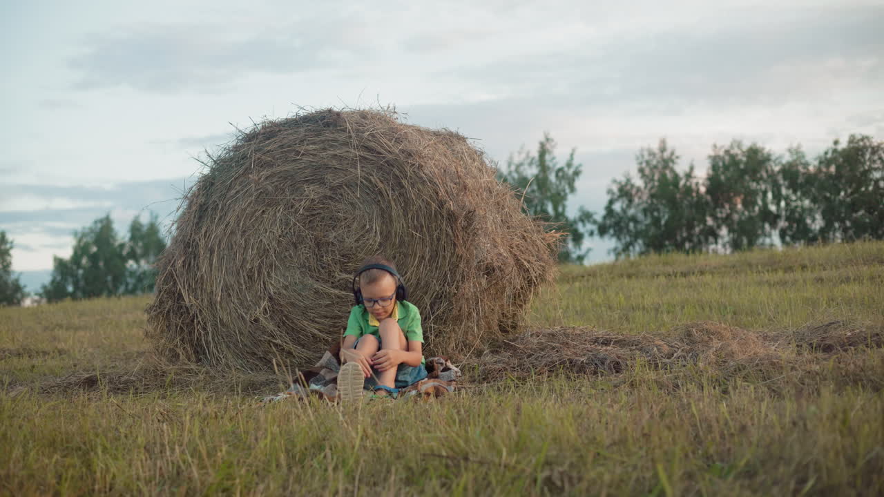 Ungt barn justerer hovedtelefoner, mens det sidder med krydsede ben foran en høballe, omgivet af vidtstrakt landbrugsland under åben himmel, og nyder et fredfyldt øjeblik i naturen.