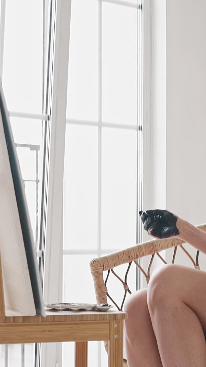Disappointed woman artist with dirty hands throws brush and leans in rattan armchair near wooden easel with bad picture in studio slow motion