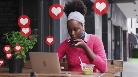 animación de los iconos del corazón de las redes sociales sobre una mujer afroamericana con computadora portátil y teléfono inteligente en un café