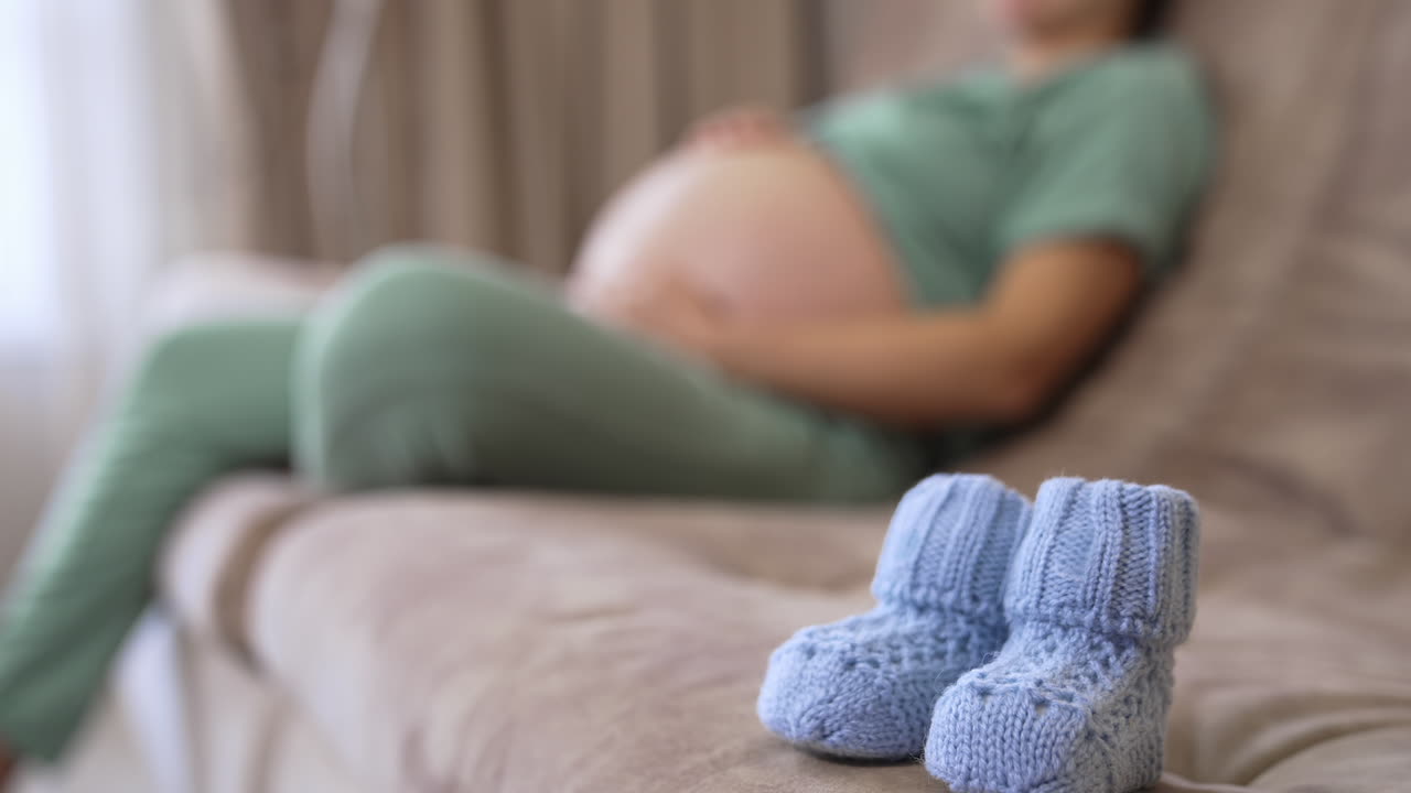 Beautiful tiny knitted socks for future child. Baby socks on blurred background of a living-room with a pregnant woman sitting on a couch and stroking her stomach.
