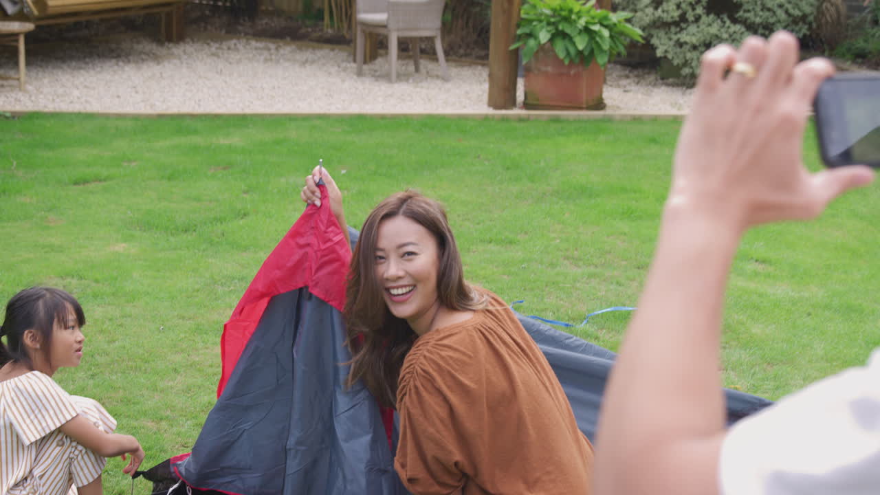 아시아인 아버지 가 집 의 정원 에 텐트 를 세우는 동안 휴대 전화 로 사진 을 찍고 있다