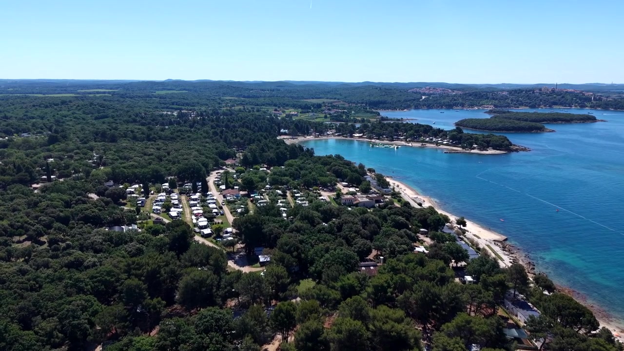 Campeggio ombreggiato da pini si estende lungo la costa di una baia croata, con acqua calma e turchese, spiaggia sabbiosa, isole distanti e città costiera sotto il sole splendente dell'estate. Croazia. Ampia ripresa aerea con drone