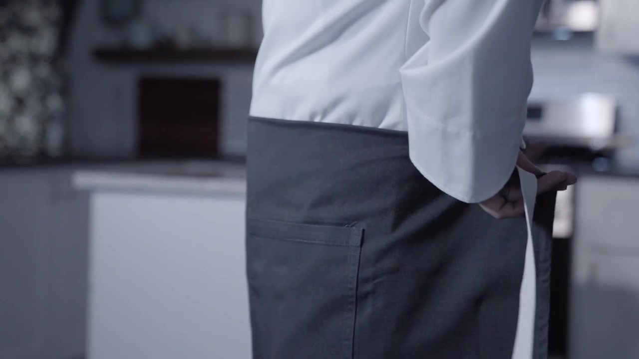Un'inquadratura laterale statica cattura uno chef maschio bianco che indossa il suo grembiule e si prepara a lavorare in cucina. Questo video evidenzia l'importanza della preparazione in cucina e dell'abbigliamento professionale