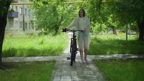 woman with blonde hair wearing raincoat strolls beside bicycle on paved path lined with lush greenery, background features trees, grass, residential building, and yellow pole