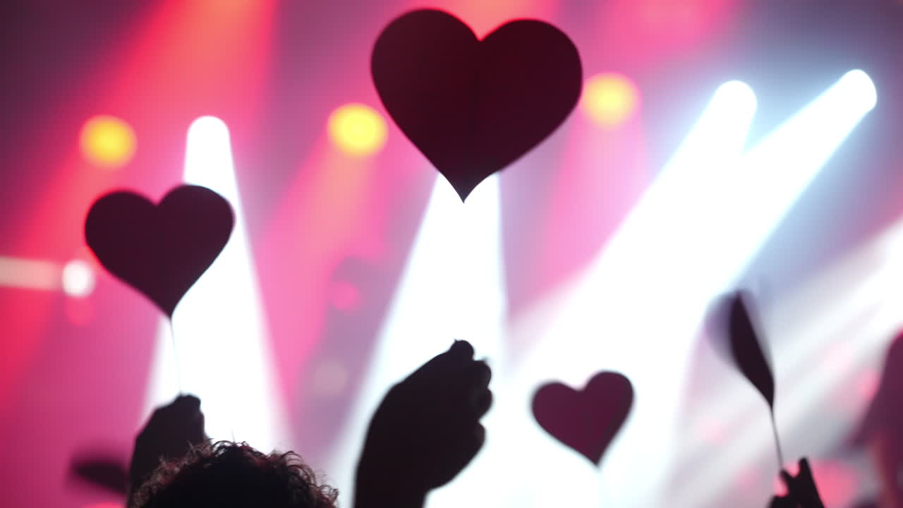 Hands holding heart-shaped signs at a vibrant concert with colorful lights and energetic atmosphere
