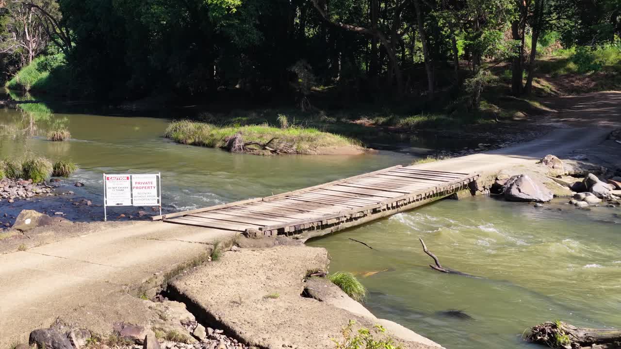 A serene stream flows through a lush forest, with a rustic bridge under bright daylight, creating a peaceful atmosphere