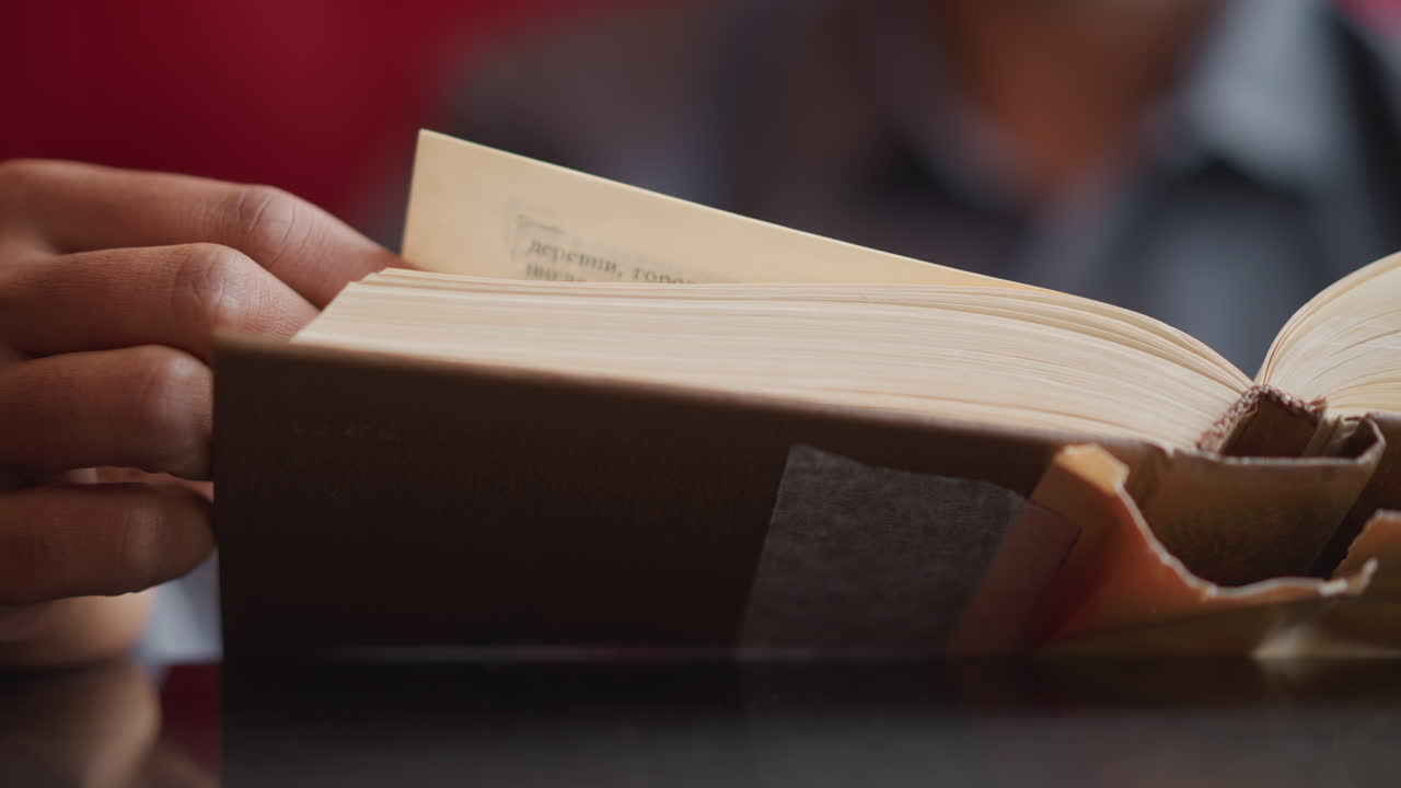 Close-up of person turning page of thick hardcover book while sitting at desk, hand mid-motion, wearing shirt, natural daylight reflecting off smooth surface, showing calm study moment or quiet research focus