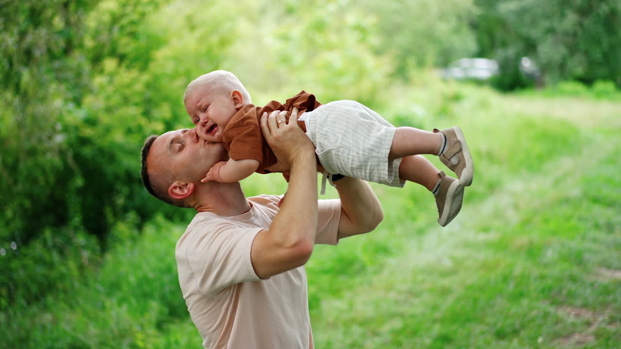 Happy father is holding his little son above the head. Kid starts to cry and dad kisses him on the cheek. Nature at backdrop.