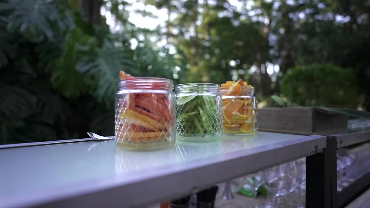 Foto reveladora de três potes de vidro contendo frutas cítricas frescas fatiadas e hortelã para um bar de coquetéis em um evento de casamento ao ar livre com um pano de fundo de jardim natural