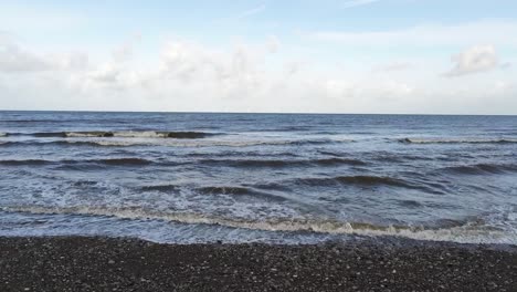 Dolly-aerial-across-ocean-waves-slashing-stony-beach-as-birds-fly-through-scene