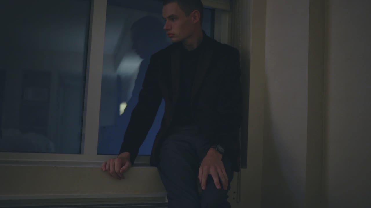 슬픈 청년은 유리창을 향해 걸어가서 밖을 바라보며 모퉁이에 앉아 있다 - 중간 클로즈업 샷