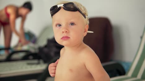 portrait of a beautiful toddler with goggles sitting by the pool and looking in the camera waiting for his mother to come