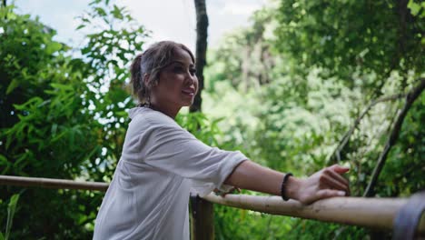 Woman-smiles-brightly-as-she-leans-on-a-wooden-railing,-surrounded-by-dense-forest-foliage