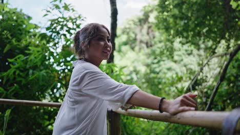 mujer sonríe brillantemente mientras se apoya en una barandilla de madera, rodeada de denso follaje del bosque