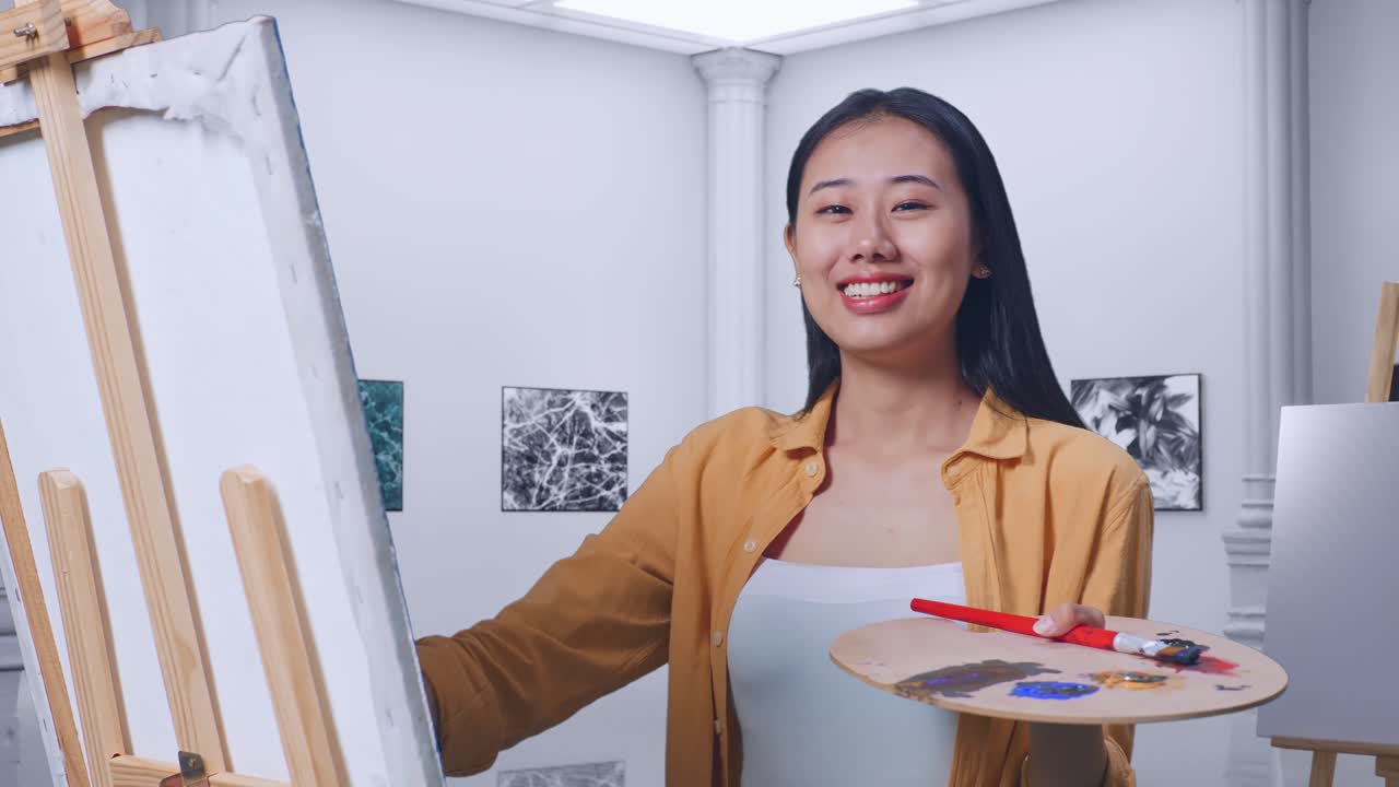아시아 여성 예술가 가 갤러리에서 오일 페인트와 으로 캔버스에 그림을 그리는 동안 미소 짓고 카메라에 은 몸짓을 보여줍니다.
