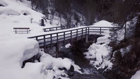 Aerial-view-of-a-small-bridge-over-a-creek-in-a-snowy-valley-in-the-alps,-Kleinwalsertal,Austria-Drone-is-lifting-up