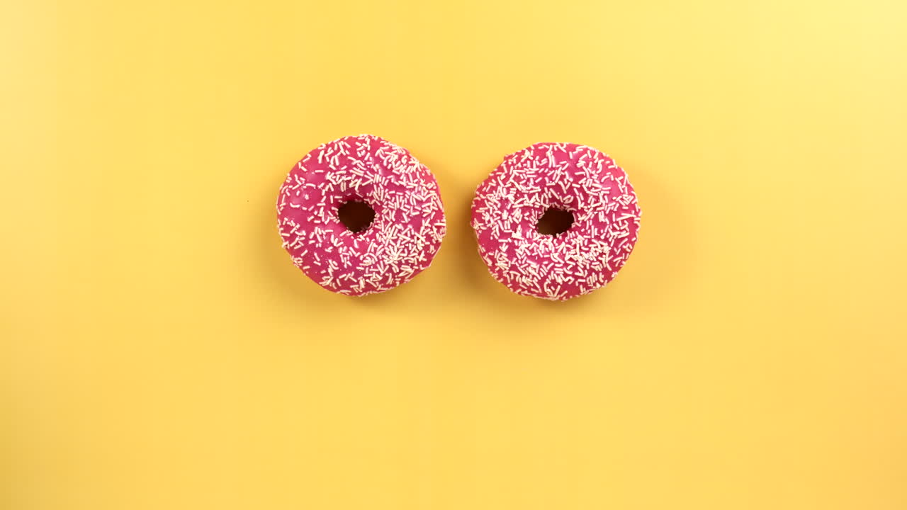 Woman take pink colorful donut on light yellow background. Female Hand grab delicious donut. Pink glazed with sprinkles. Filmed in Prores, Topview, Slowmotion