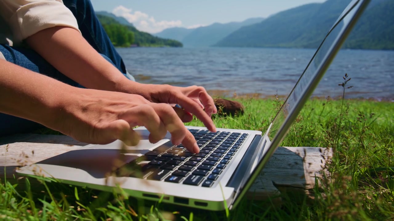 Person taster på bærbar computer ved siden af rolig sø, grønne bakker bagved. Hænder bevæger sig hurtigt, fokuseret på skærmen. Fredelig udendørs arbejdsscene, naturligt lys. Ideel til fjernarbejde, rejser eller digitale nomadetemaer.
