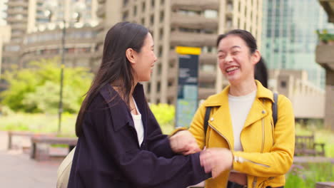 Two-Smiling-Young-Female-Friends-Meet-Talking-As-They-Walk-Outdoors-Along-City-Street-Together-In-Real-Time