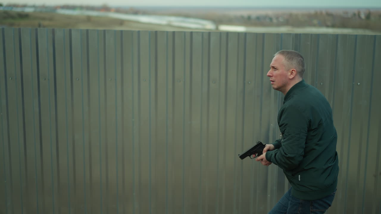 A close view of a man in a green jacket holding a handgun and moving cautiously along a metal fence in an industrial area