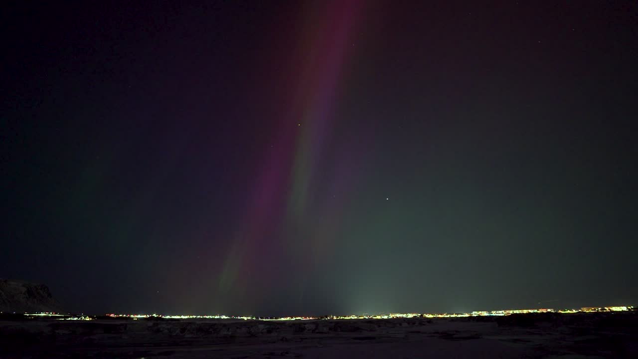 Green and purple northern lights over Icelandic city at night. Time lapse wide shot. Aurora Borealis with magical sky. Snowy winter day.