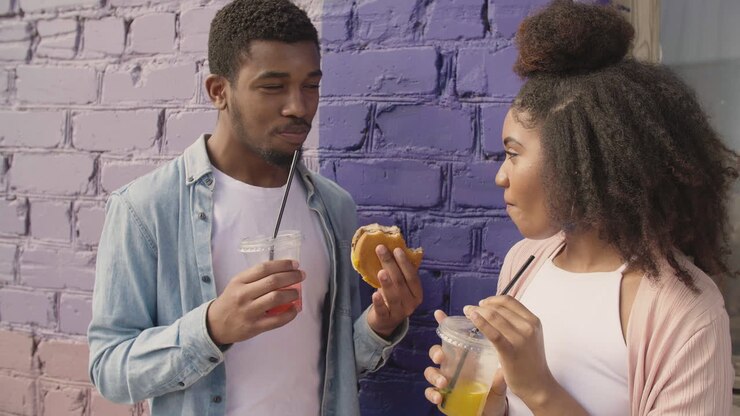 Young Couple Sharing A Tasty Hamburger And Drinking A Cold Drink Together, While Chatting Leaning Against A Colourful Brick Wall 1