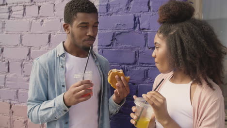 young couple sharing a tasty hamburger and drinking a cold drink together, while chatting leaning against a colourful brick wall 1