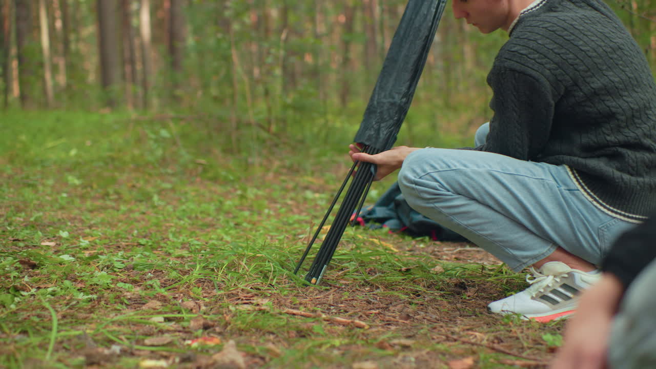 Close up of man squatting on forest ground carefully pulling tent pole from black bag during camping preparation while another person observes from nearby in peaceful woodland setting