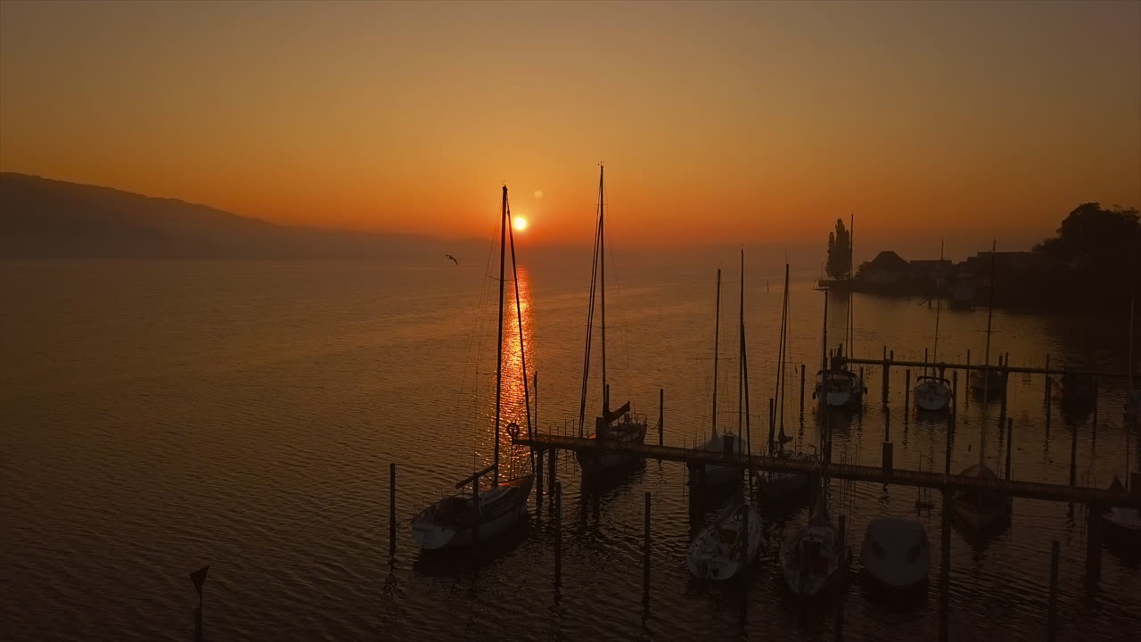 Calm lakeside harbor at Lake Constance at sunrise with an orange sky and boats whose masts reflect in the water. The sun is just above the horizon, casting a glowing path across the lake.