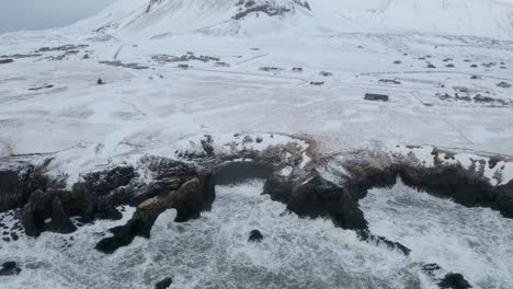 Aerial-tilt-down-shot-of-snowy-Snæfellsjökull-Volcano-and-waves-of-ocean-with-steep-dark-cliffs-in-foreground---Visiting-Iceland-by-Winter