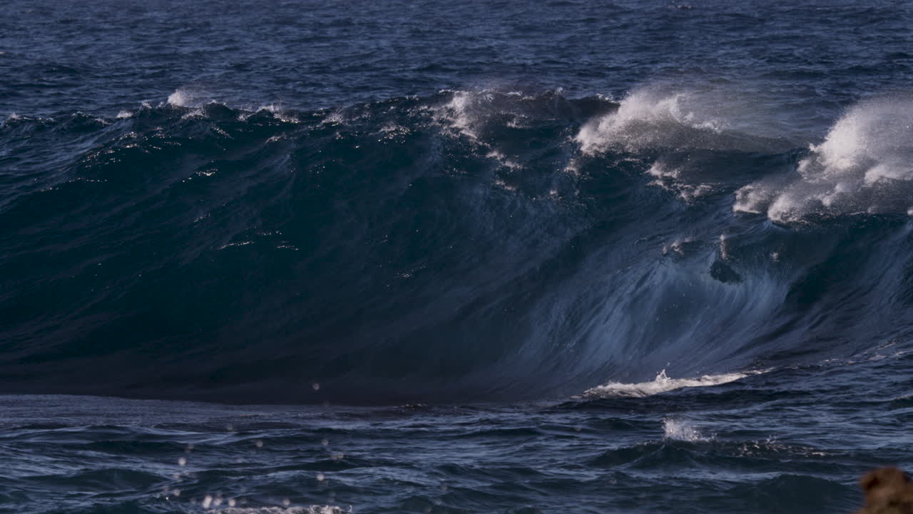 A thick, heavy wave breaking in slow motion onto a shallow reef as a strong offshore wind blows spray high into the air causing flashes of rainbow colour