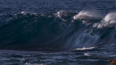 a thick, heavy wave breaking in slow motion onto a shallow reef as a strong offshore wind blows spray high into the air causing flashes of rainbow colour