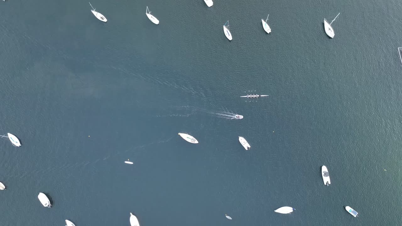Wide overhead tracking aerial shot of row boats heading towards the shore past docked boats