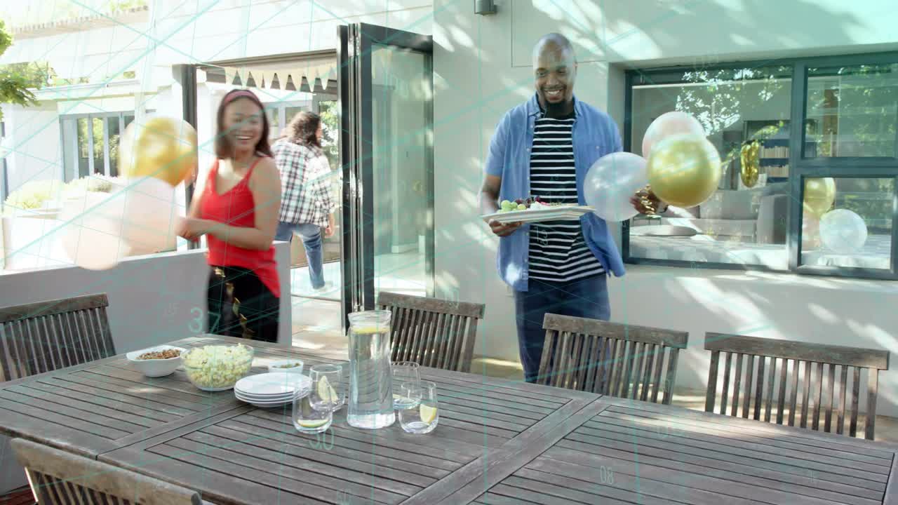 Man in plaid arranging plates on outdoor table, friends entering door with balloons preparing meal