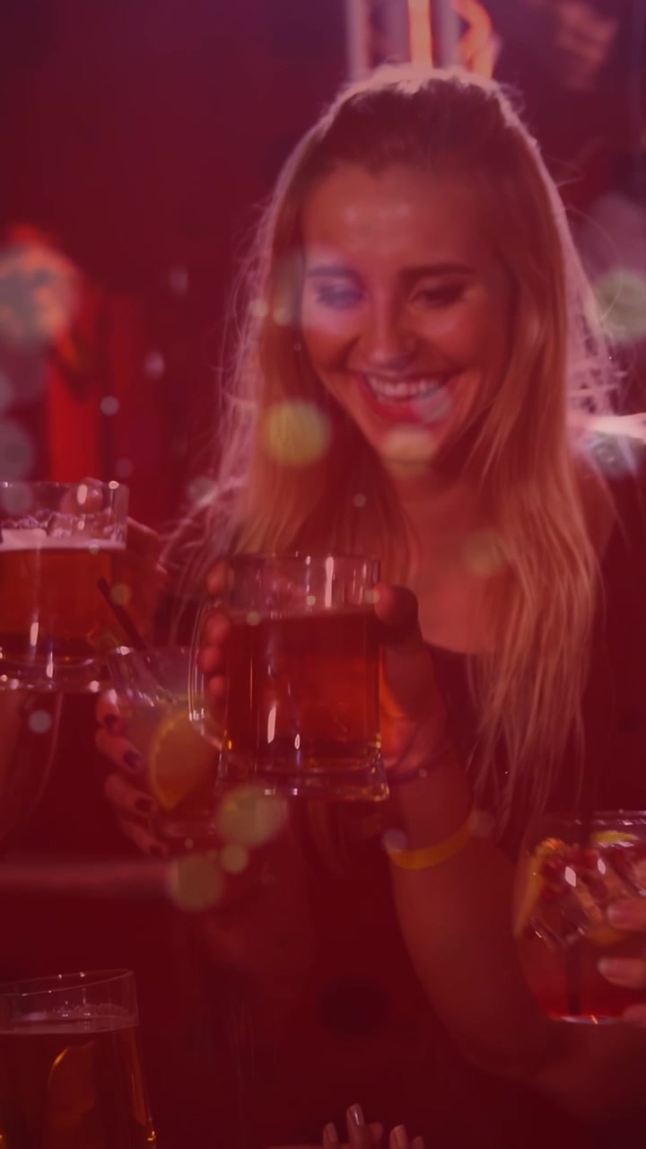 Video verticale: Donna che solleva entrambi i drink dopo il brindisi dell'amica, con effetto bokeh che si sovrappone ai drink in un bar.