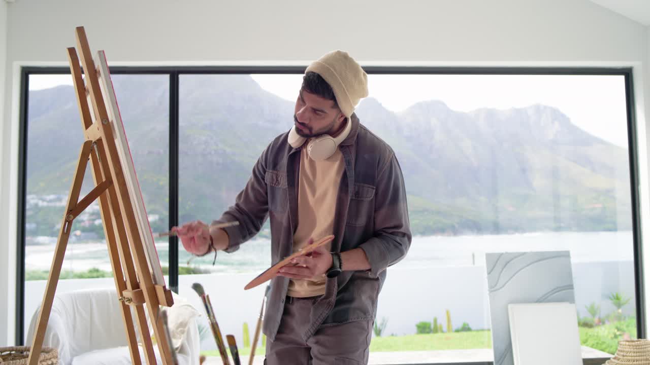 Indian male painter referencing view, dipping brush and painting canvas at home studio with palette