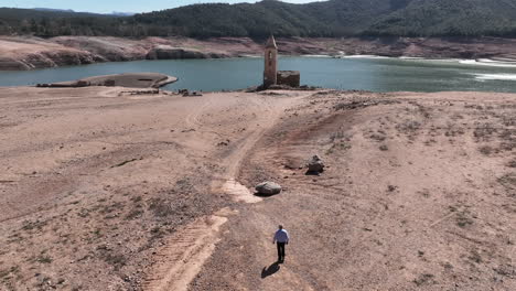 Confident-man-walking-towards-church-tower-of-drying-lake-coastline,-aerial-view