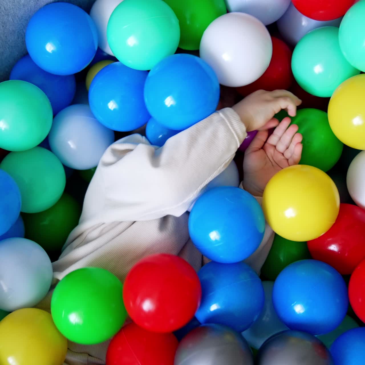 Baby boy lies hiding in the colorful balls in a dry pool. Toddler turns and sits in the basin taking one red ball in hands. Top view