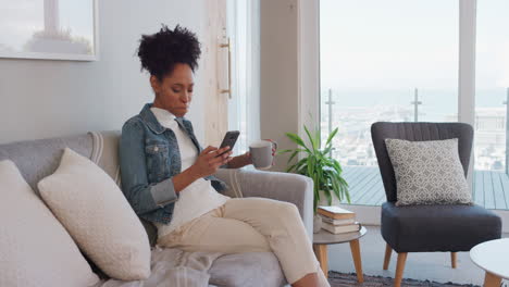 Mujer-Joven-Feliz-Usando-Un-Teléfono-Inteligente-Navegando-En-Línea-Leyendo-Mensajes-De-Redes-Sociales-Disfrutando-De-Un-Estilo-De-Vida-Cómodo-Bebiendo-Café-Relajándose-En-El-Sofá-En-Casa